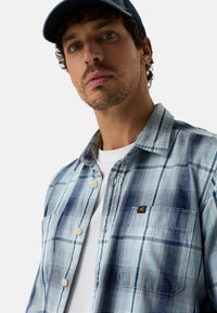 Blau kariertes Hemd mit durchgehendem Knopfverschluss, weißen und marineblauen Linien, einer Brusttasche und einem Logodetail auf dem Stoff.