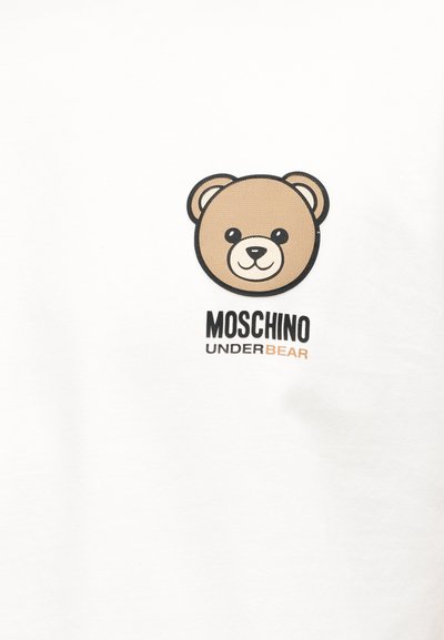 Χαμογελαστό καρτούν πρόσωπο αρκούδας πάνω από το κείμενο «MOSCHINO UNDERBEAR» σε απλό λευκό φόντο, στο κέντρο ενός υφάσματος.