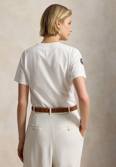 Ξανθιά γυναίκα με κοντά μαλλιά που φοράει λευκό κοντομάνικο πουκάμισο με μαύρο λογότυπο στο μανίκι, λευκό παντελόνι και καφέ πλεκτή ζώνη.