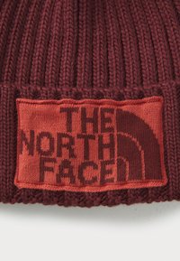 Strikket burgunderrød hue med et tekstureret ribmønster, prydet med et kontrastfuldt orange patch, der viser "THE NORTH FACE" logoet.