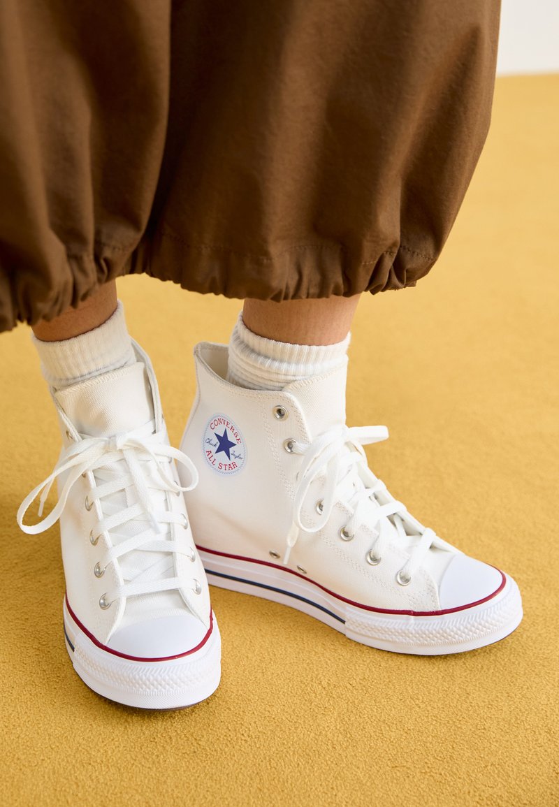 Zapatillas altas de lona blancas con suela de goma, que cuentan con ojales plateados, un ribete rojo y azul, y un logo circular en el tobillo.