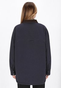 Persona con capelli lunghi che indossa una giacca larga di un blu navy scuro con polsini abbottonati e un colletto marrone, in piedi su uno sfondo bianco.