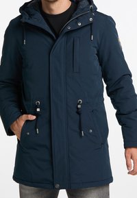 Uomo che indossa un cappotto invernale blu navy con cappuccio, cerniera, bottoni a pressione, lacci e tasche frontali, abbinato a jeans grigi.
