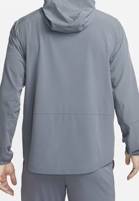 Grijze jas met een capuchon, voorzien van een gladde textuur en een rechte snit. Opvallende naaddetails bij de schouders en onderrug.