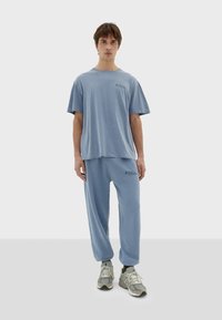 Světle modré tričko s krátkým rukávem a odpovídající tepláky z měkkého materiálu. Výrazný detail loga na tričku. Sportovní obuv dokončuje outfit.