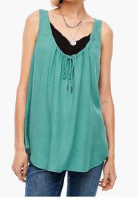 Blusa sin mangas en color verde azulado con escote redondo, detalle fruncido y cordón ajustable. Llevada sobre una camiseta negra, combinada con vaqueros azules.