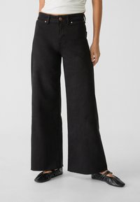 Wijdbenige zwarte jeans van stevig denim, met een hoge taille, zichtbare stiksels en een rauwe zoom. Gecombineerd met zwarte platte schoenen.