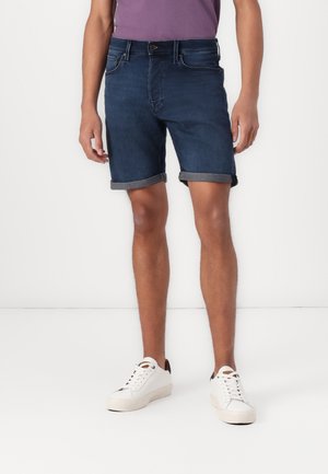 Moški, oblečen v temno modre zavihane jeans kratke hlače, bele superge in vijolično majico, stoji pred enobarvnim belim ozadjem.