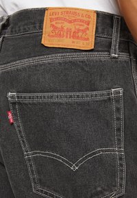 Poche arrière et étiquette à la taille d’un jean noir Levi’s porté par une personne vêtue d’une chemise blanche, montrant le patch de la marque et l’étiquette rouge Levi’s.