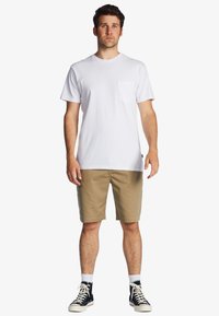 T-shirt bianca in cotone con taschino sul lato sinistro, abbinata a pantaloni beige. Indossata con sneakers alte bianco e nero. Design semplice e casual.