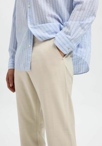 Camisa de botones a rayas azul clara con mangas largas y pantalones beige ajustados, que presentan una textura suave y un detalle de bolsillo lateral.