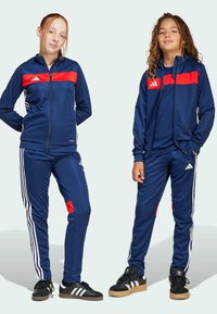 Twee kinderen dragen bijpassende marineblauwe sportoutfits met rode strepen, staan zelfverzekerd met de handen op de heupen tegen een lichte achtergrond.