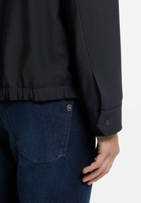 Dark navy jacket with a smooth fabric, elastic waistband, and buttoned cuffs, paired with blue denim pants featuring a back pocket.