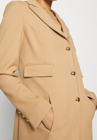 Cappotto beige su misura con dettagli in bottoni dorati, colletto a revers e tasche laterali. Realizzato in un materiale morbido e texturizzato, con una vestibilità strutturata.