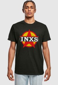 Sort bomulds t-shirt med et rundt rødt og gult logo med en stjerne og teksten "INXS" i fed hvidt på tværs af midten.