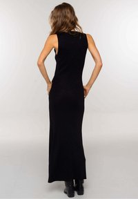 Femme aux cheveux ondulés portant une robe maxi noire sans manches et des bottes noires, se tenant les mains sur les hanches, de dos.
