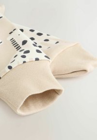 Beżowy sweter z czarnymi i brązowymi kropkami, prążkowanymi mankietami oraz miękką teksturą. Projekt zawiera zabawne motywy zwierzęce.