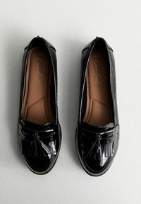 Mocasines de charol negro con detalle de borlas en la parte frontal, plantilla acolchada en beige y diseño de punta redondeada suave. Clásicos y sencillos.