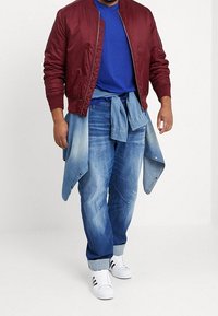 Burgunderfärgad bomberjacka över en blå t-shirt, ljusblå jeans med uppvikta fållar, och vita sneakers med svarta ränder. Jeansskjorta knuten i midjan.