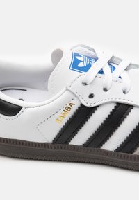 Sapatilha de couro branco com riscas pretas e texto dourado "SAMBA". Apresenta um logótipo azul da Adidas, atacadores brancos e sola de borracha preta texturizada.