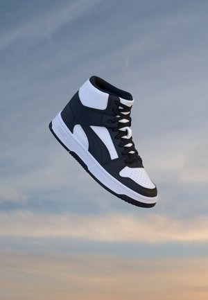 Basket montante noir et blanc avec lacets flottant devant un ciel nuageux au coucher du soleil.