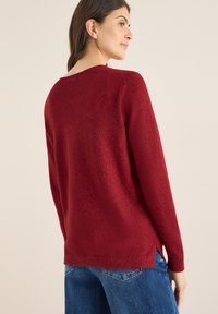 Czerwona swetrowa bluzka z długimi rękawami, okrągłym dekoltem i bocznymi rozcięciami. Miękka tekstura z delikatnym połyskiem. Zestawiona z niebieskimi dżinsami.