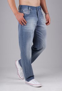 Man wearing light blue faded jeans and white sneakers, standing on a gray studio floor against a gray background.