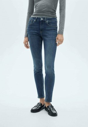 Personne portant un jean bleu ajusté, un haut gris à manches longues, des mocassins noirs et des bagues aux doigts, debout devant un fond clair.