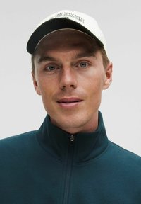 Tmavomodrý zipsový kabát s vysokým golierom, skombinovaný s bielou čiapkou s zeleným textom, na svetlom neutrálom pozadí.