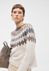 Κρέμα πουλόβερ με γεωμετρικό πλέγμα σε καφέ και μπλε, στρογγυλή λαιμόκοψη και υφή. Το μοντέλο κρατά μια γκρίζα σουέντ τσάντα.