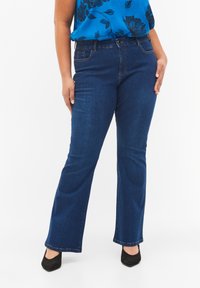 Jeans acampanados de denim azul con una cintura de altura media, costuras contrastantes y un cierre frontal con botón. El material parece suave y duradero.