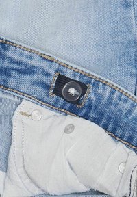 Jean en denim bleu clair avec une étiquette noire visible, un bouton transparent et des fermetures en argent. Doublure intérieure blanche au niveau des poches.