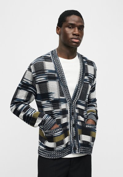 Νεαρός άντρας που φοράει μαύρη, λευκή, μπλε και ελιά μοτίβο πλεκτή ζακέτα πάνω από λευκό πουκάμισο με τα χέρια στις τσέπες της ζακέτας.