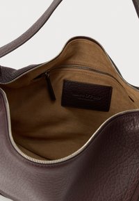 Odpeta temno rjava usnjena torbica s bež semiš notranjostjo, z žepom z zadrgo znotraj in vidno šivano znamko na oblogi.