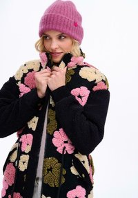 Čierna fleecová bunda s kvetinovým vzorom, na ktorej sú ružové, žlté a zelené kvety. Má vysoký golier, zips a rebrované čierne manžety. Tiež je zobrazená svetloružová pletená čiapka.
