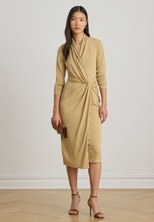 Femeie purtând o rochie midi bej cu stil învelit și mâneci trei-sferturi, ținând o poșetă maro și purtând sandale maro cu toc înalt.