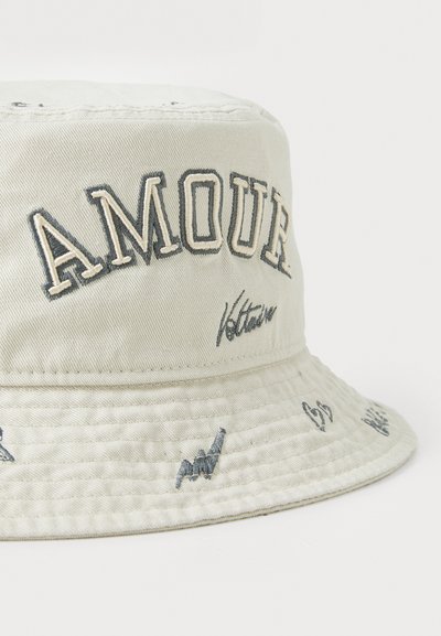 Kapa s širokim obodom od svijetlosmeđeg pamuka s izvezenim natpisom "AMOUR" u tamnoplavoj boji, koja ima razne male izvezene uzorke oko oboda.