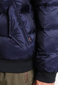 Granatowa kurtka puchowa z pikowanym wzorem, boczną kieszenią, elastycznymi mankietami i ściągaczem u dołu dla większego ciepła i wygody.