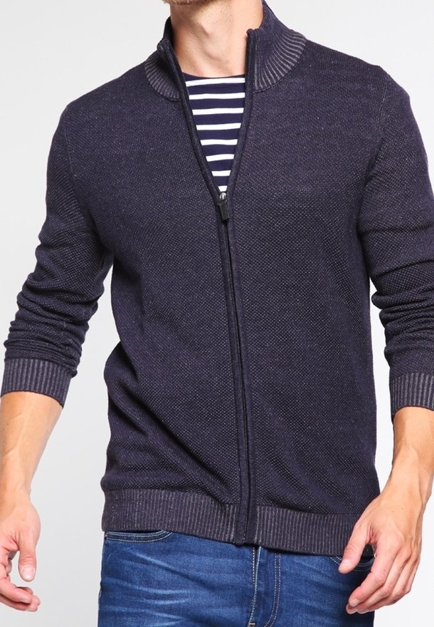 Granatowy sweter zapinany na zamek z teksturowanym wzorem dzianiny, z prążkowanym kołnierzem i mankietami, noszony na paskowanym podkoszulku, w zestawieniu z niebieskimi dżinsami.
