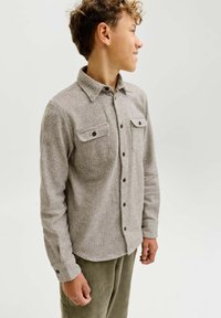 Camicia button-up grigia con un tessuto testurizzato, due tasche frontali e bottoni neri, abbinata a pantaloni in cotone a coste verde. Colletto liscio, vestibilità rilassata.