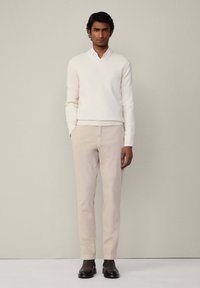 Hellbeige gestrickter Pullover mit V-Ausschnitt, kombiniert mit hellbraunen Chinos und schwarzen Lederstiefeln. Schlichtes, elegantes Design ohne Muster.