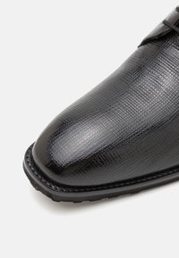 Chaussure de ville en cuir noir avec une surface texturée, bout rond et semelle en caoutchouc noir. Présente des coutures discrètes le long du bord.