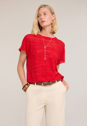 Lyshåret kvinde iført en rød kortærmet top, beige bukser med brun bælte, guldhalskæde, øreringe og armbånd, stående med hænderne i lommerne.