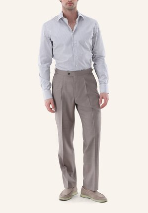 Mann trägt graue plissierte Hose, hellblau-weiß gestreiftes Langarmhemd und beige Slipper, steht vor einem neutralen Hintergrund.