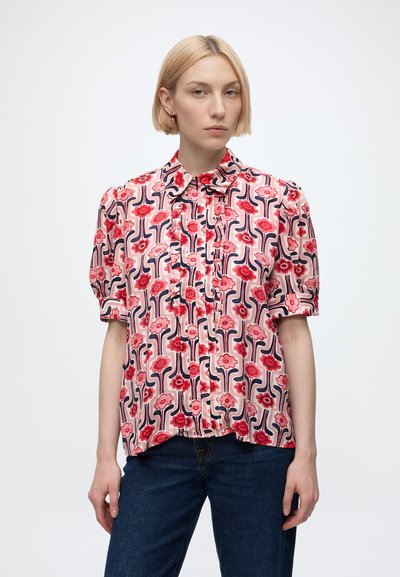 Femme aux cheveux blonds courts portant un chemisier rose à manches courtes avec des motifs floraux rouges et ondulés bleus et un jean bleu foncé.