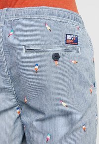 Shorts rayés bleu et blanc avec des motifs de sucettes glacées multicolores brodées. Comprend une poche arrière avec un bouton noir et une étiquette de marque.