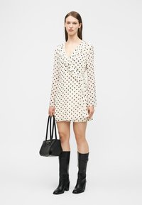 Γυναίκα που φοράει λευκό μίνι φόρεμα με V-neck και μακριά μανίκια με μαύρα σχέδια καρδιές, κρατώντας μαύρη τσάντα και φοράει μαύρες μπότες μέχρι το γόνατο.