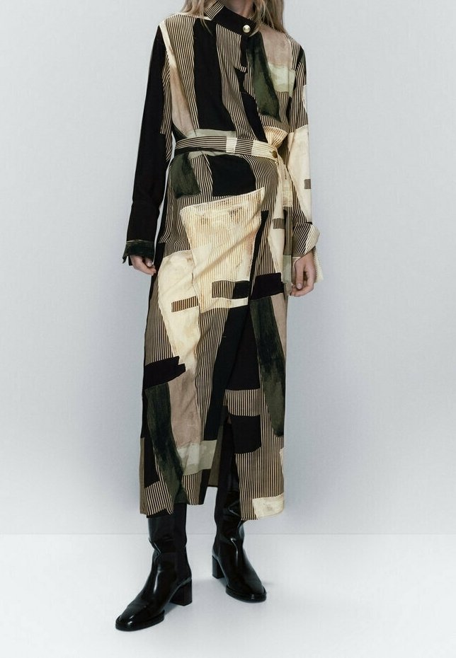 Trench-coat long à motifs avec des blocs abstraits beige, noir et vert, taille ceinturée, porté avec des bottes noires à talons sur un fond uni.