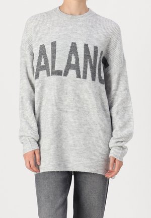 Jasnoszary sweter dzianinowy z czarnymi blokowymi literami "ALANG", noszony z ciemnoszarymi dżinsami, widok z przodu na tors, ramiona swobodnie opadające wzdłuż ciała.