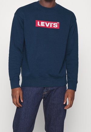 Mężczyzna w granatowej bluzie Levi's z czerwonym i białym logo, w połączeniu z ciemnoniebieskimi jeansami, stojący na jednolitym tle.